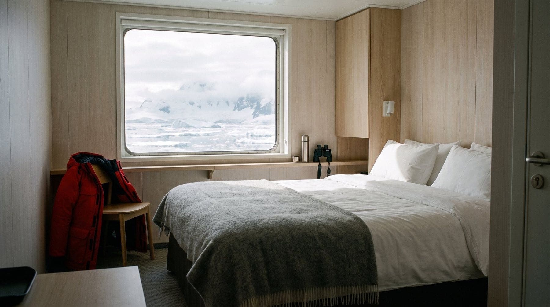 A cabin on a small expedition ship, a made bed beside a large window onto sea ice and mountains