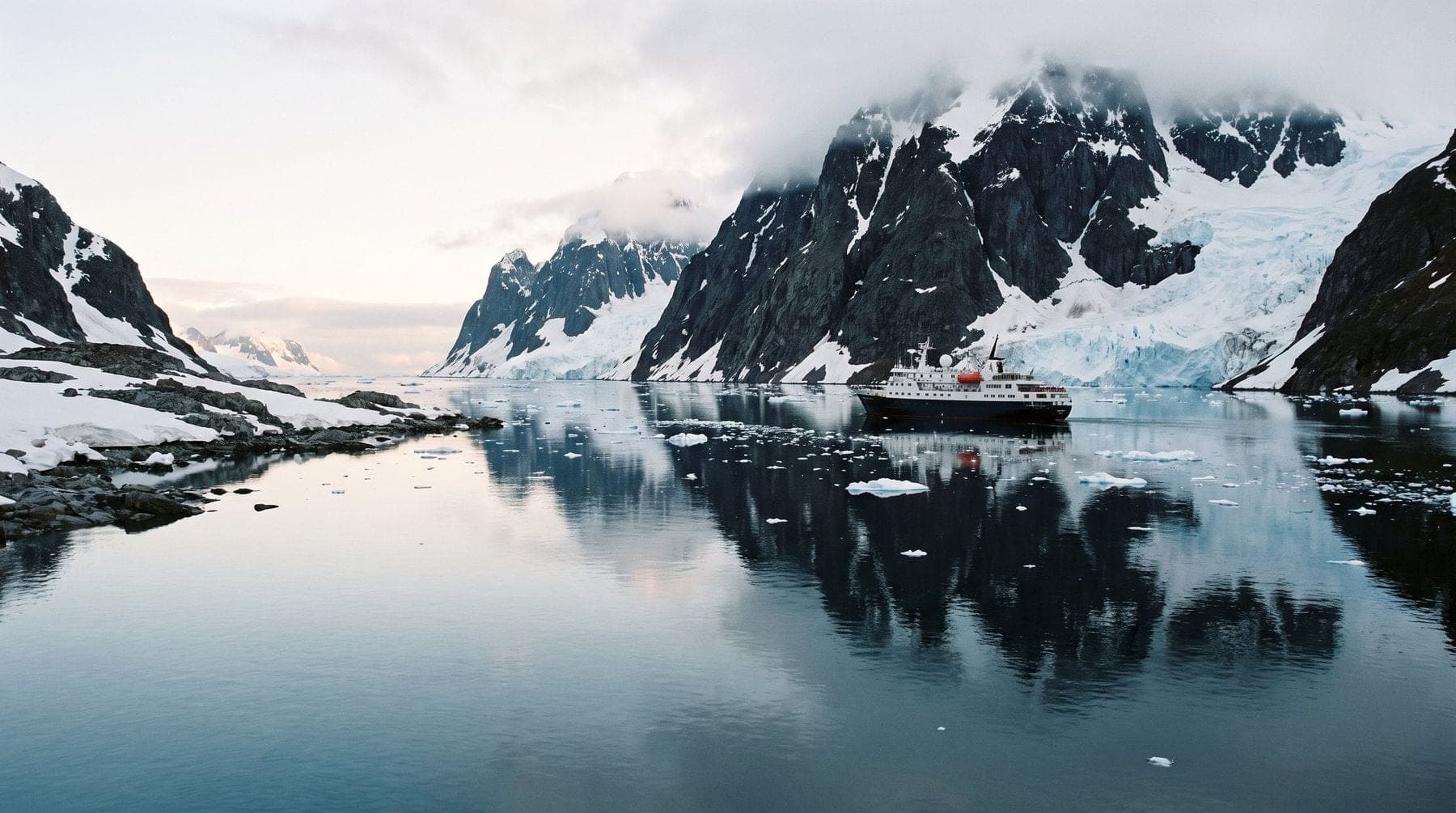 An expedition ship at rest on mirror-calm water between snow-covered mountains