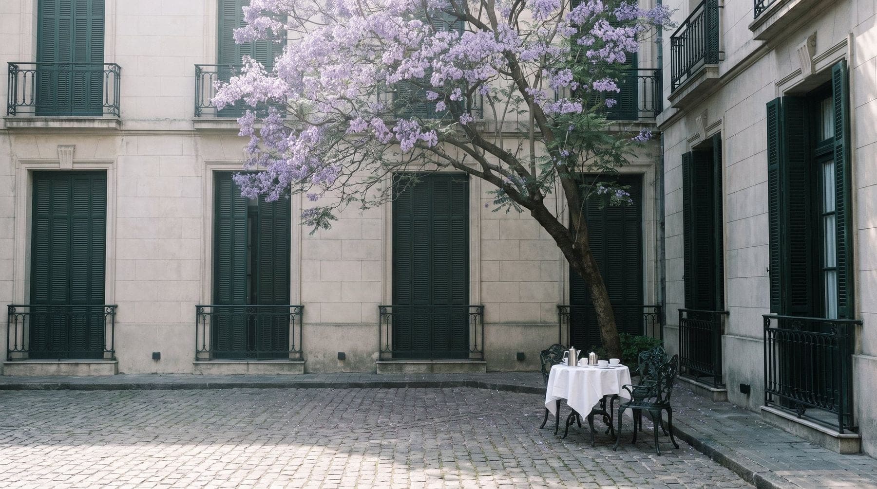 A quiet stone hotel courtyard with a jacaranda in bloom and a breakfast table for two