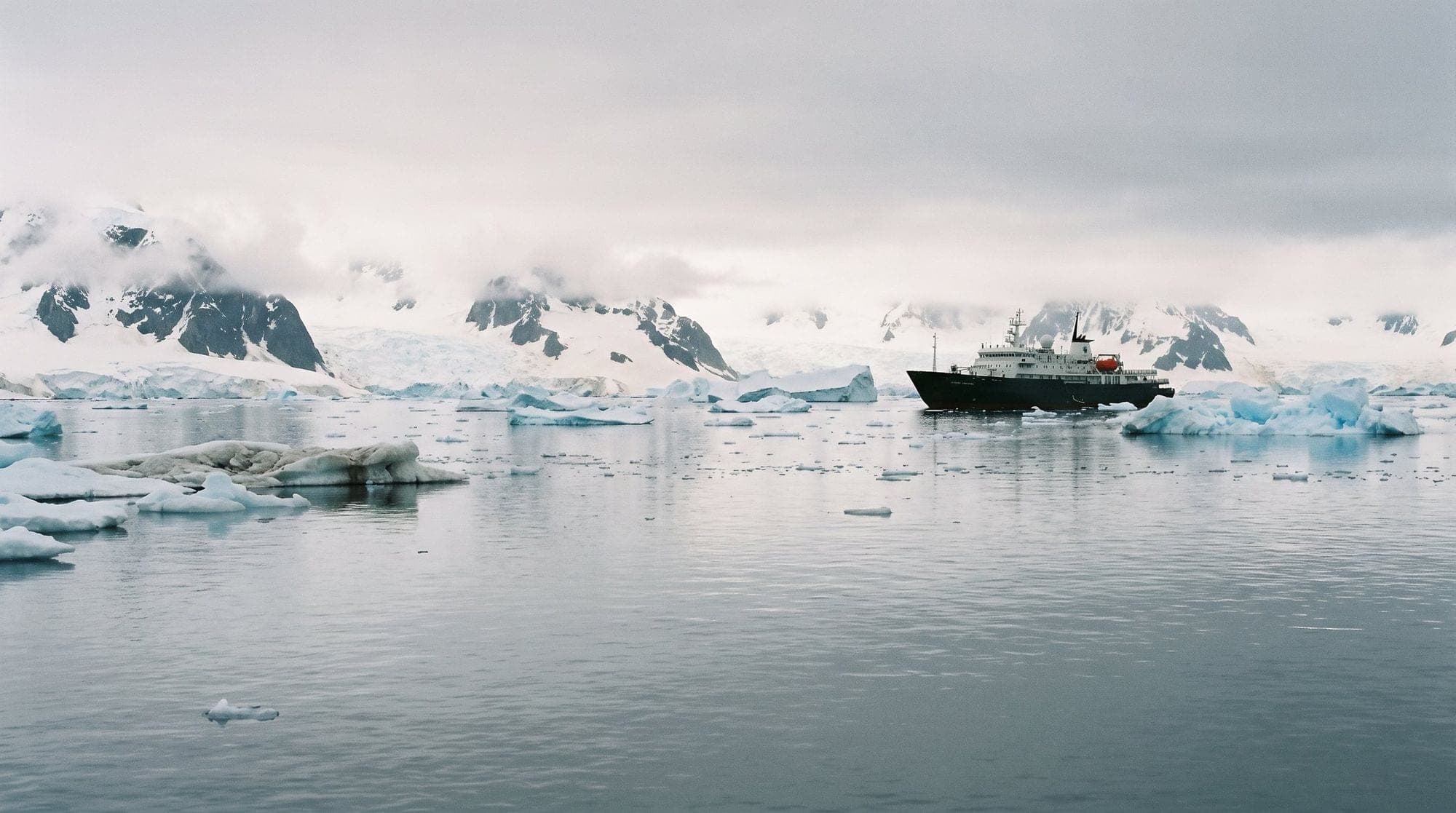 A small expedition ship among icebergs below snow-covered peaks in Antarctica