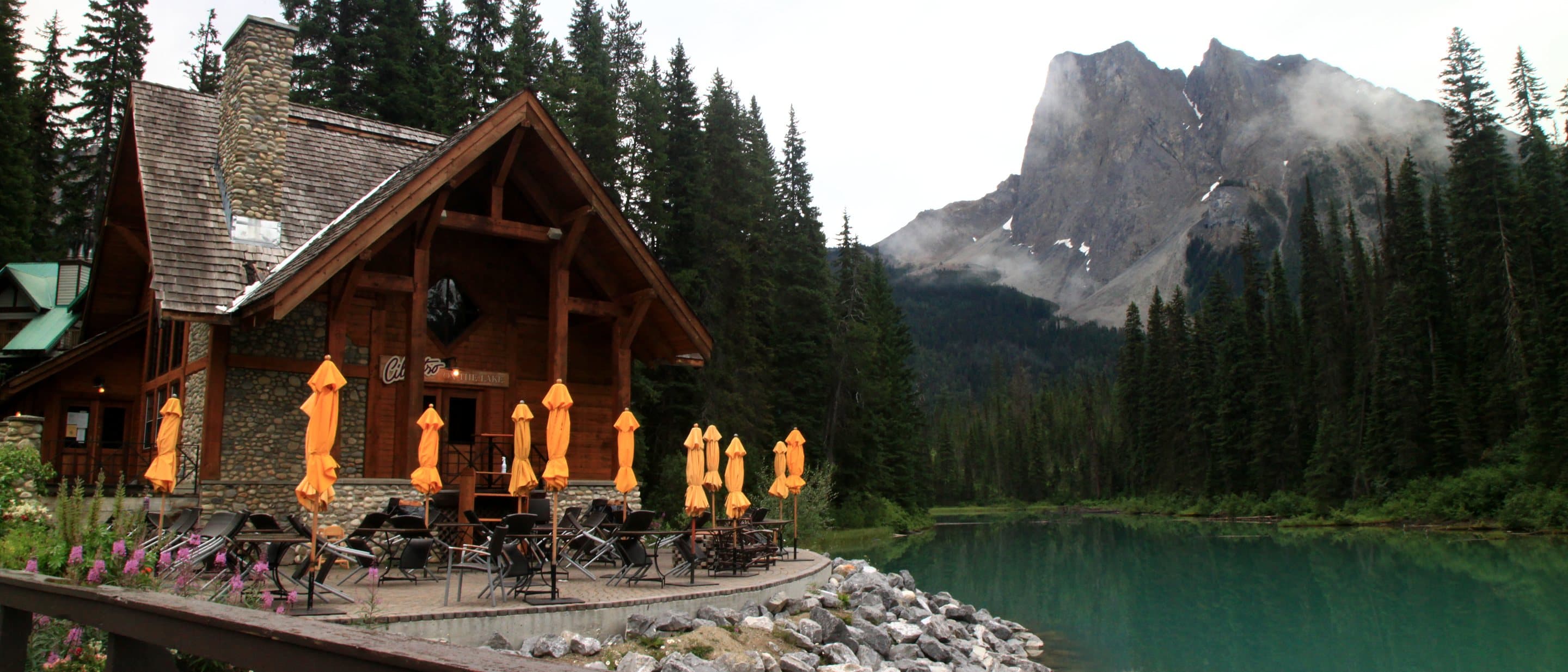 A timber lodge building with orange umbrellas on its terrace beside the turquoise water of Emerald Lake, a rocky peak behind