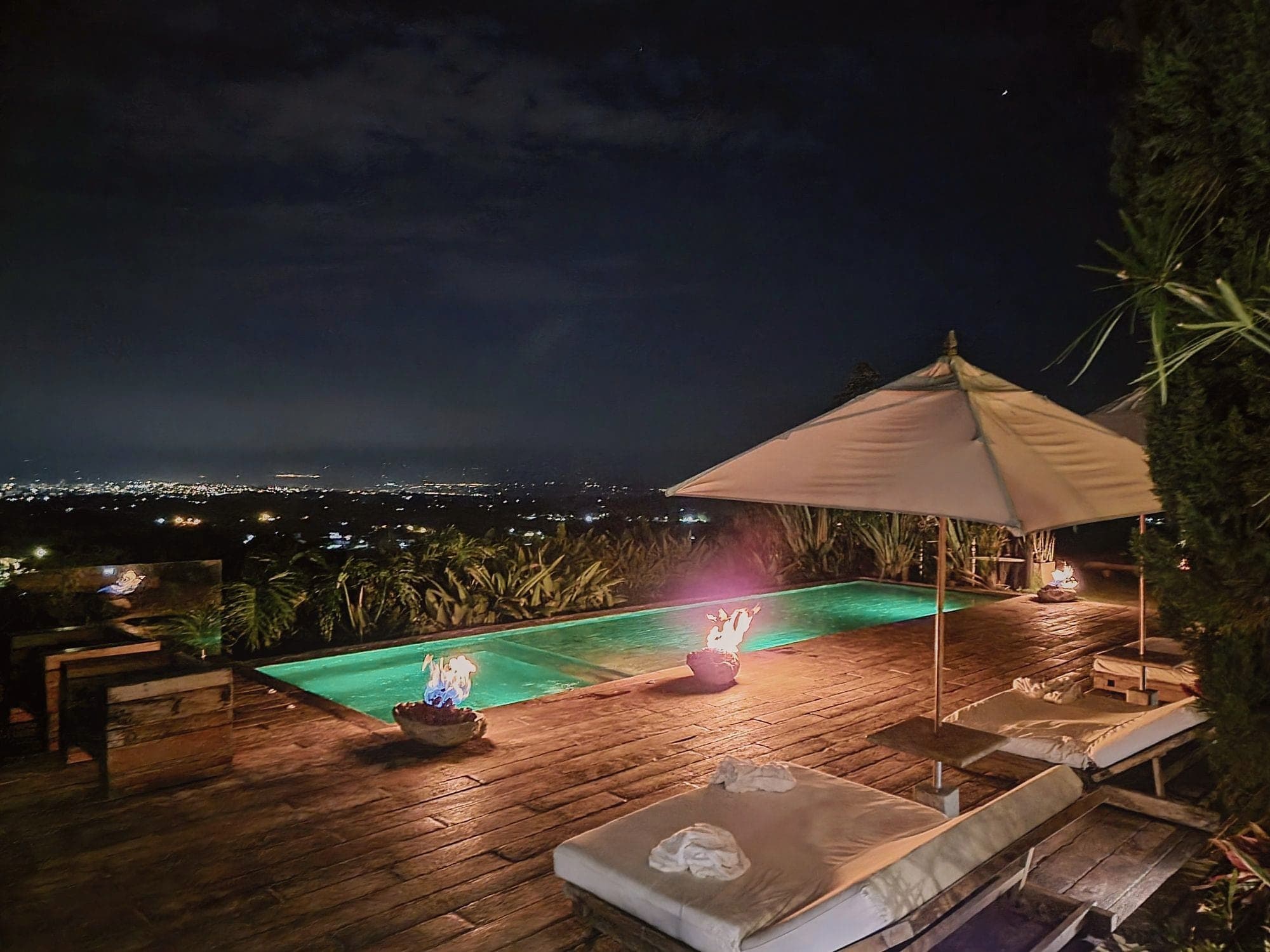 A lit plunge pool and umbrella on a suite deck at night, town lights in the valley below