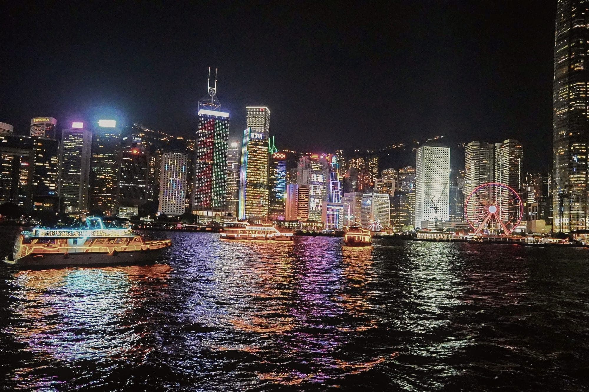 The Hong Kong skyline lit at night, reflected in Victoria Harbour, a ferry crossing in the foreground.