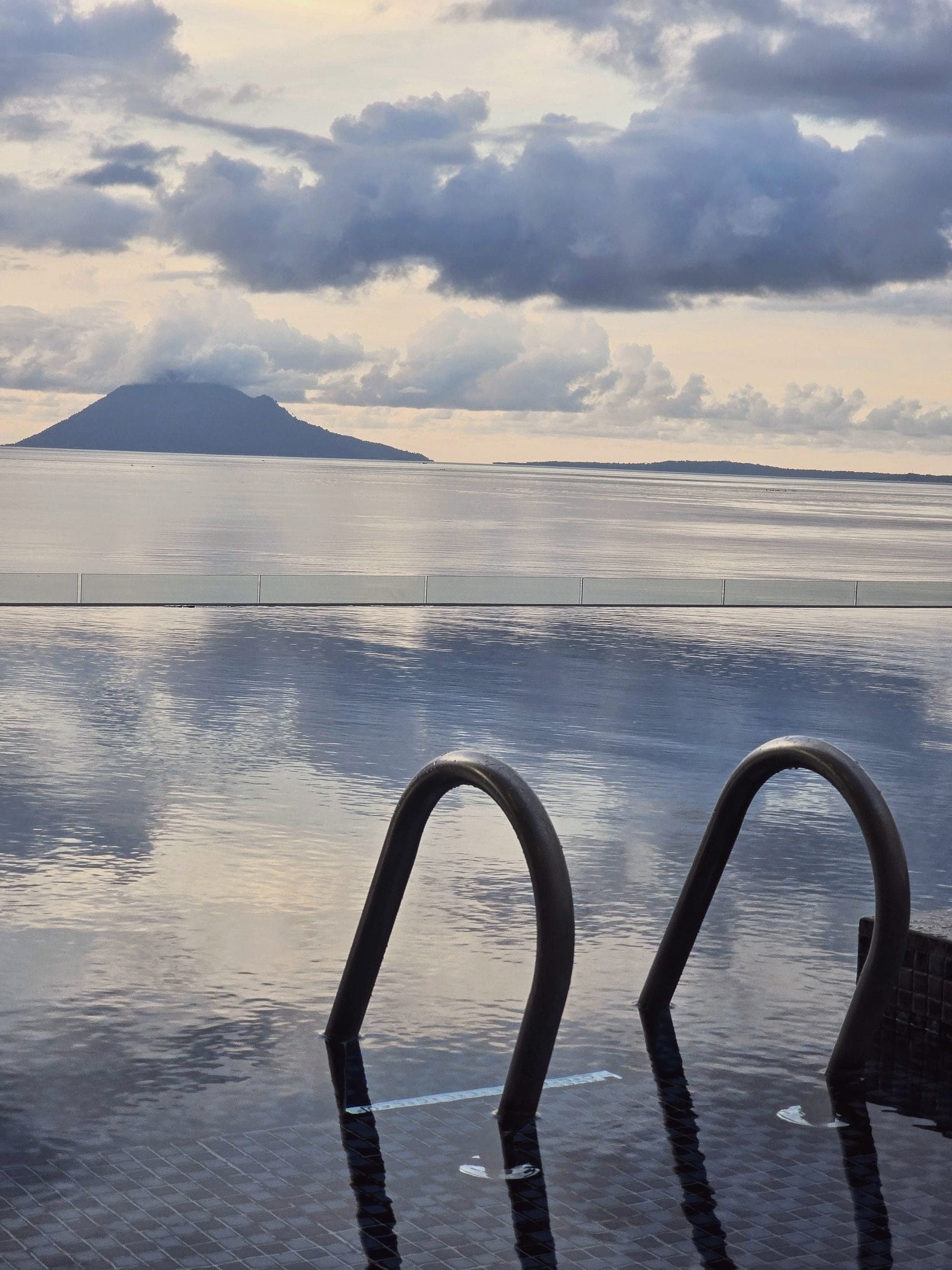 The ladder of an infinity pool looking out over a calm sea to the volcano of Manado Tua under heavy cloud, North Sulawesi, Indonesia.