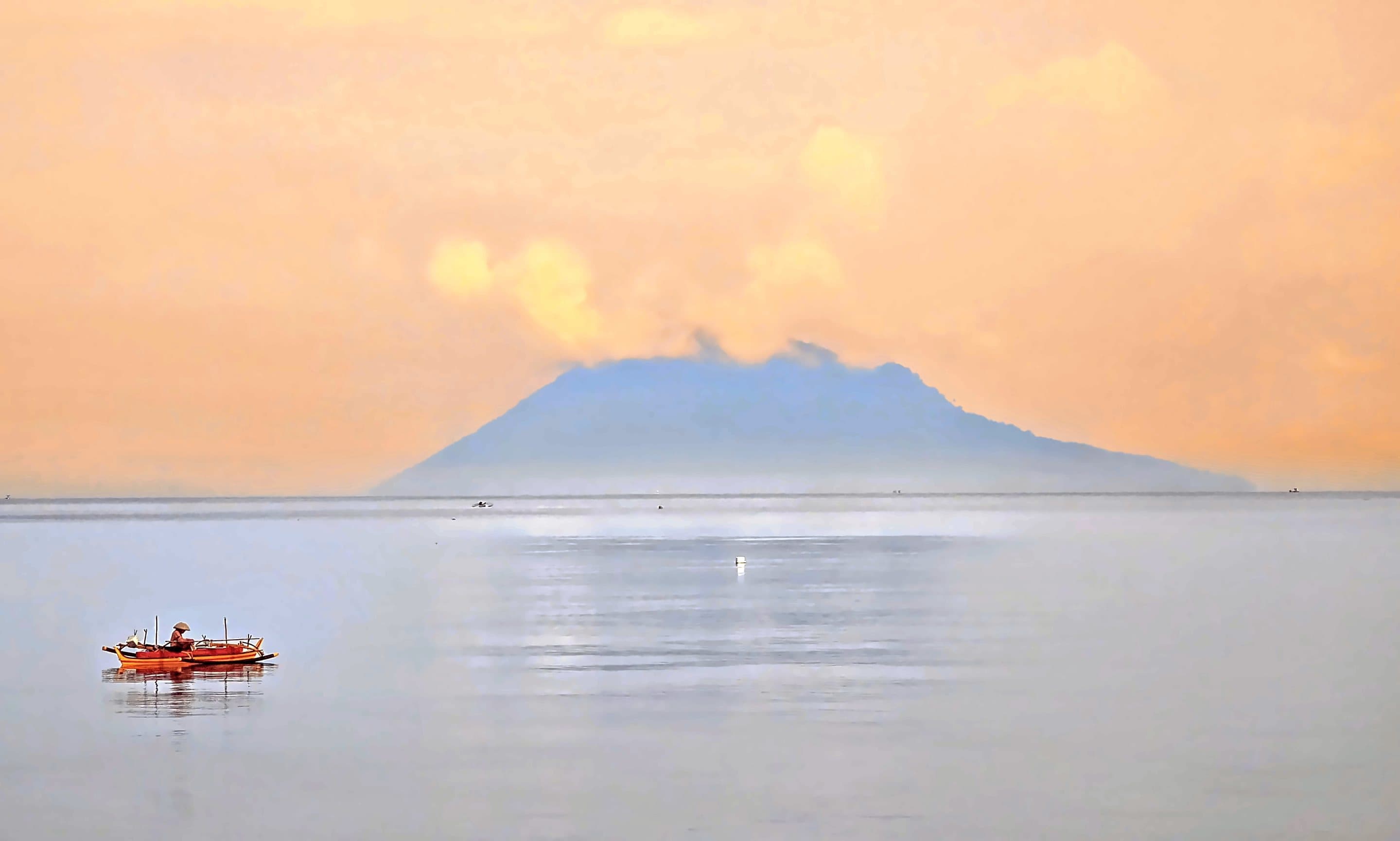 A calm, glassy sea at dawn under soft pink cloud, the volcano of Manado Tua small on the horizon, North Sulawesi, Indonesia.