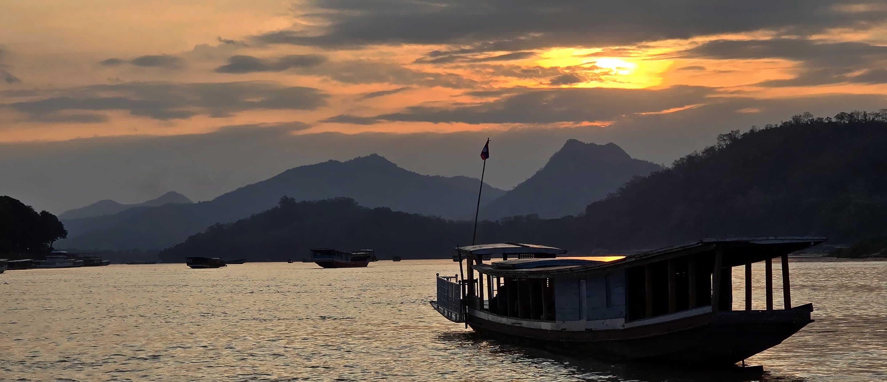 A wooden river boat moored on a wide river at sunset below mountain silhouettes, Laos