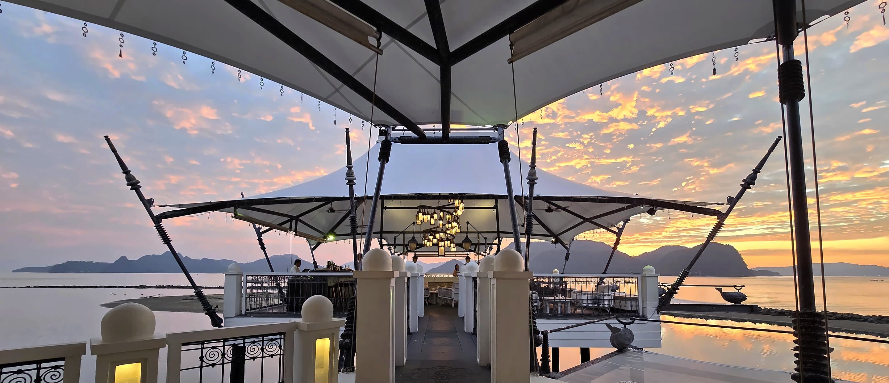 A white pier walkway leading to a tented pavilion at pink and gold sunset, islands on the horizon, Malaysia.