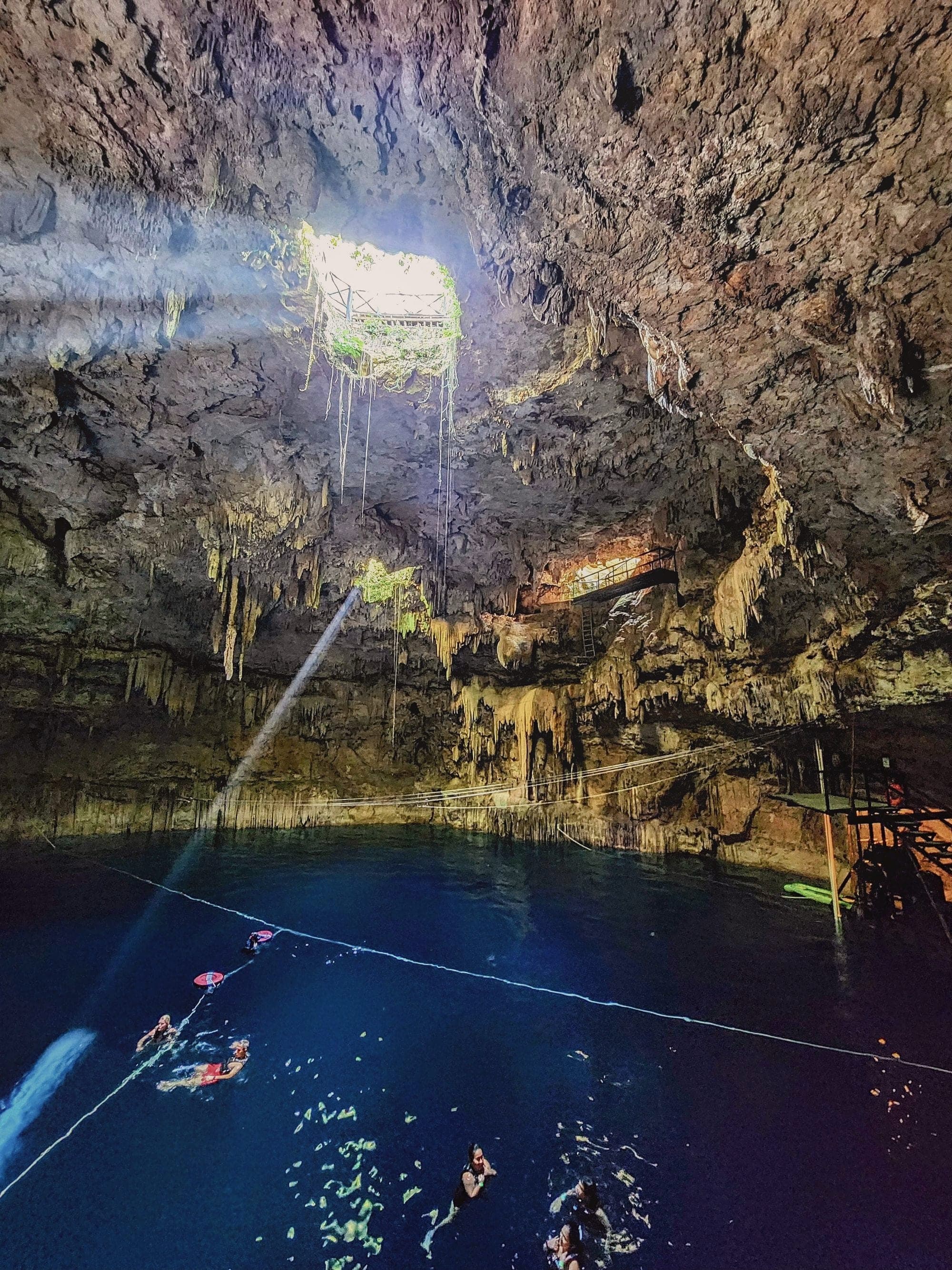 A beam of light falling through a cave roof into a cenote, swimmers below