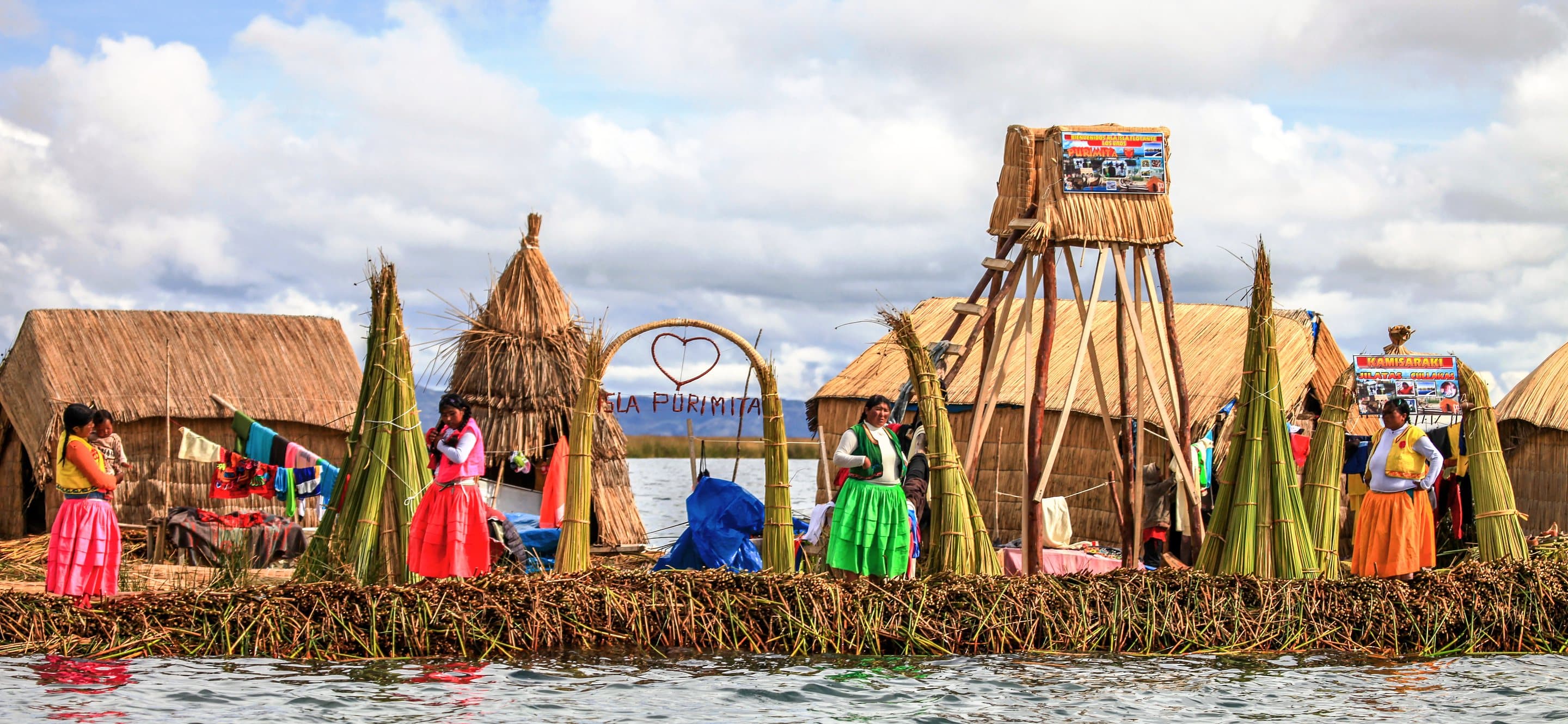 Women in bright skirts standing on a floating reed island beside reed houses and a reed watchtower