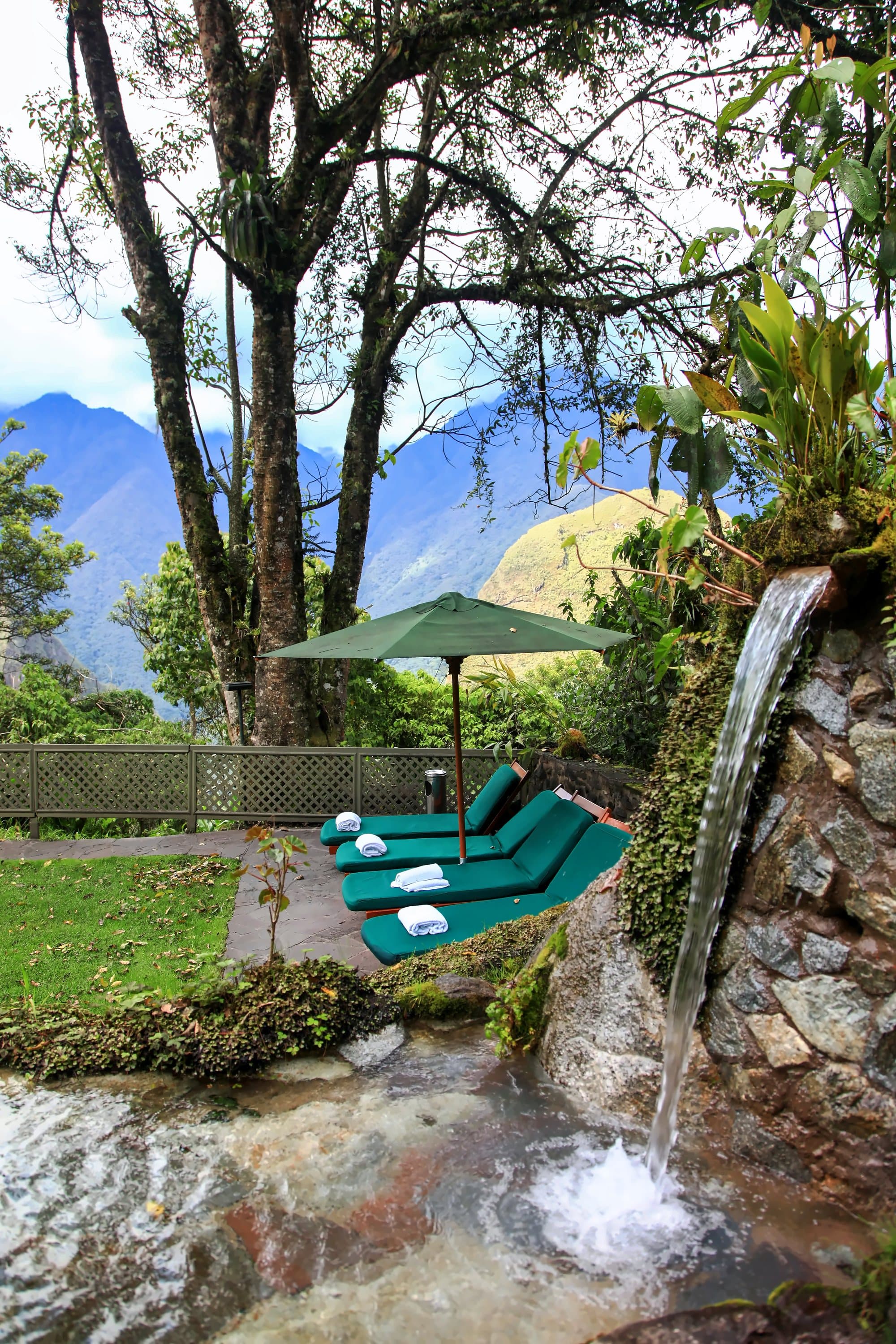 A lodge garden with a stone waterfall, loungers and an umbrella below green peaks, Machu Picchu, Peru.