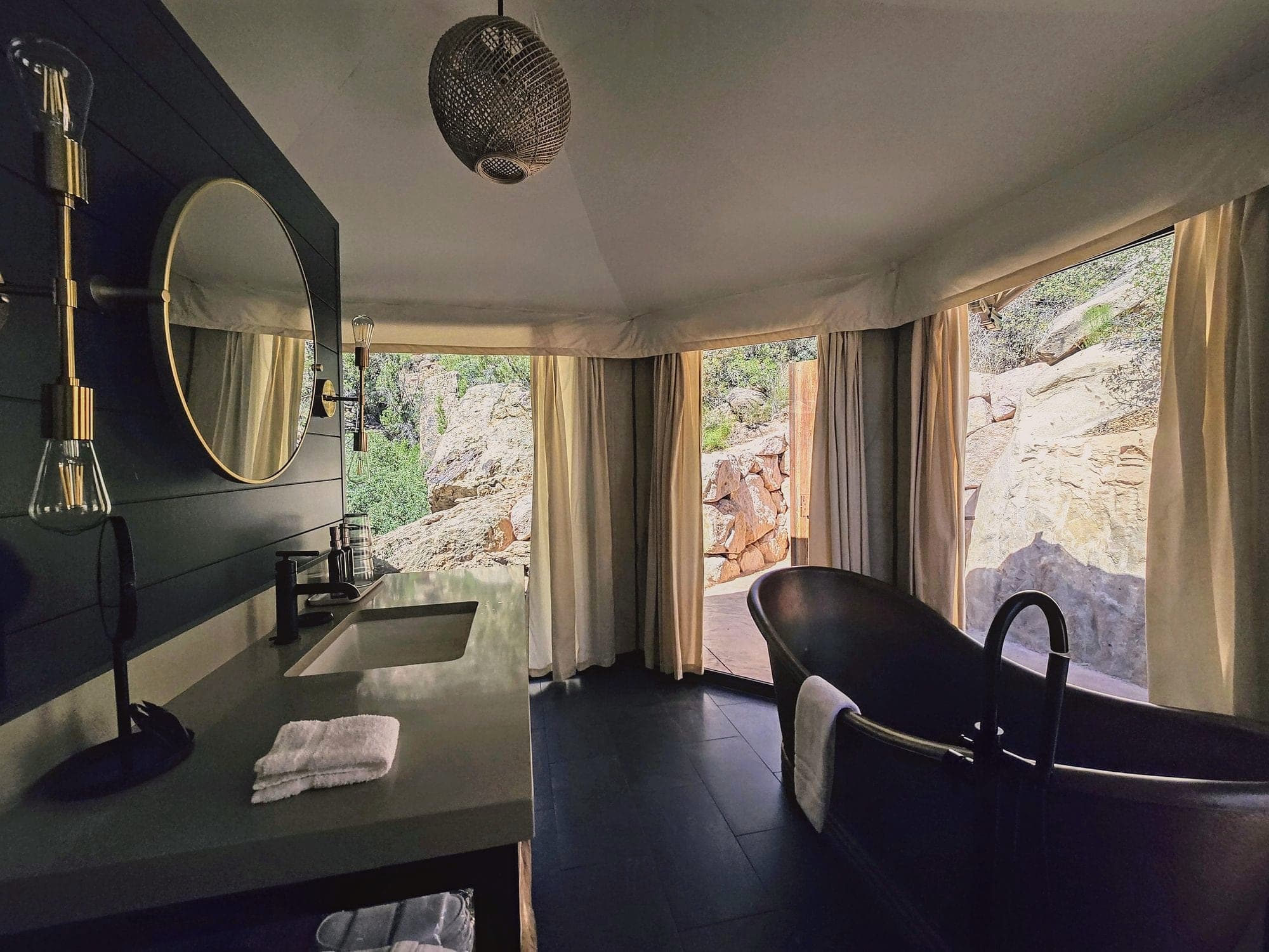 A freestanding black bathtub and vanity by tall curtained windows inside the tent