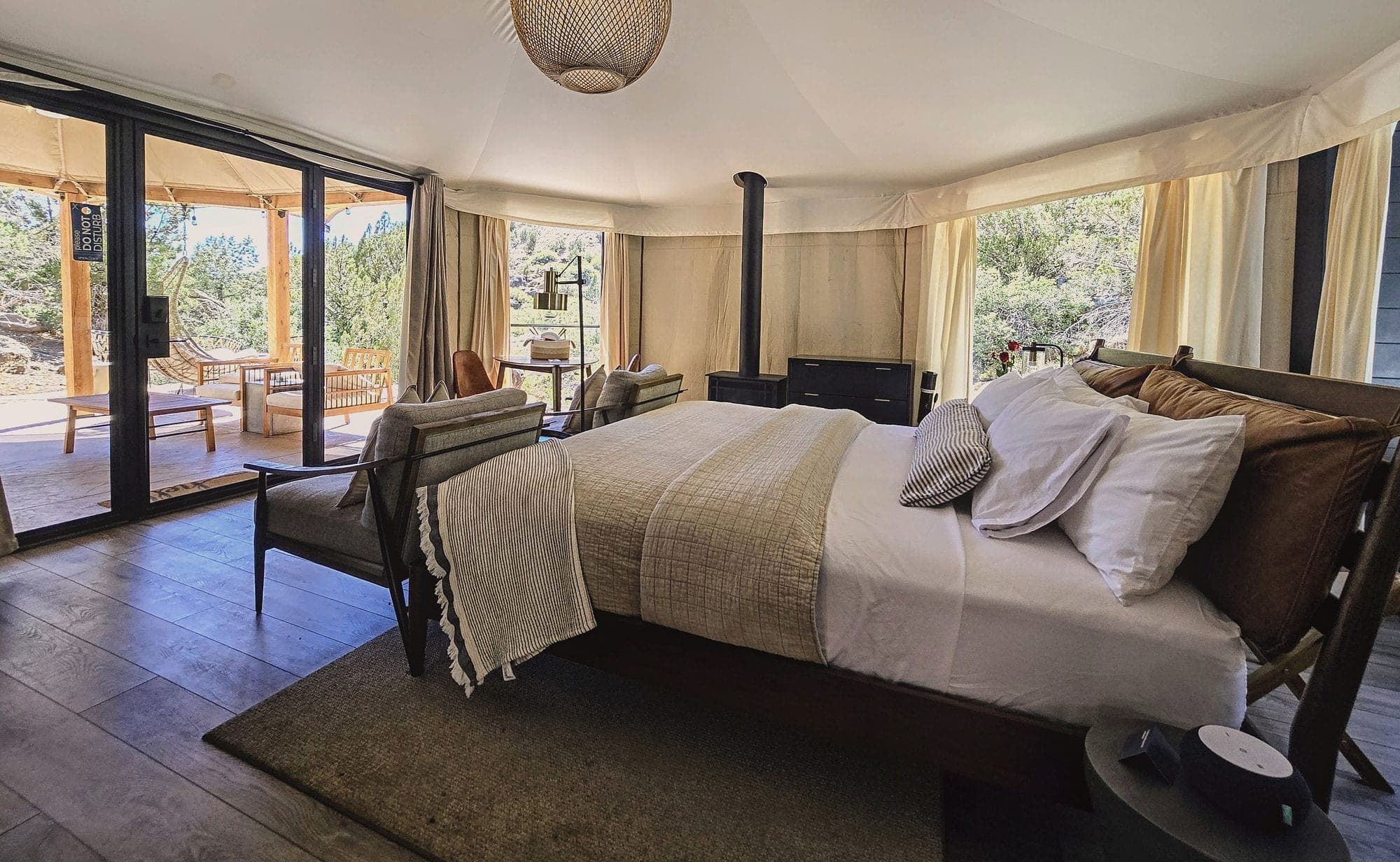 A canvas suite bedroom with a made bed, armchair and wood stove, the front open to the deck and desert scrub, near Zion, Utah.