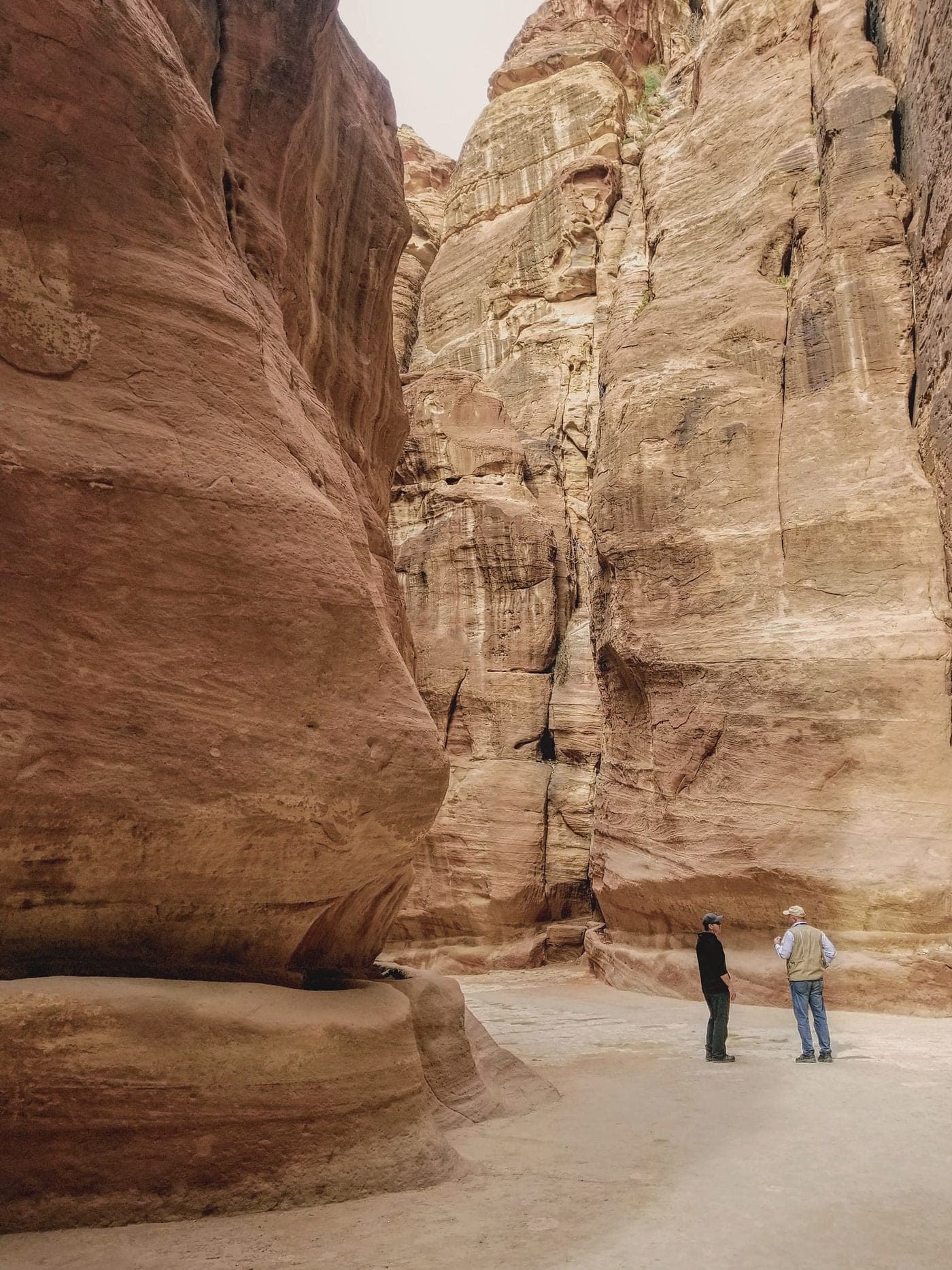 Two people small at the base of the towering walls of the Siq